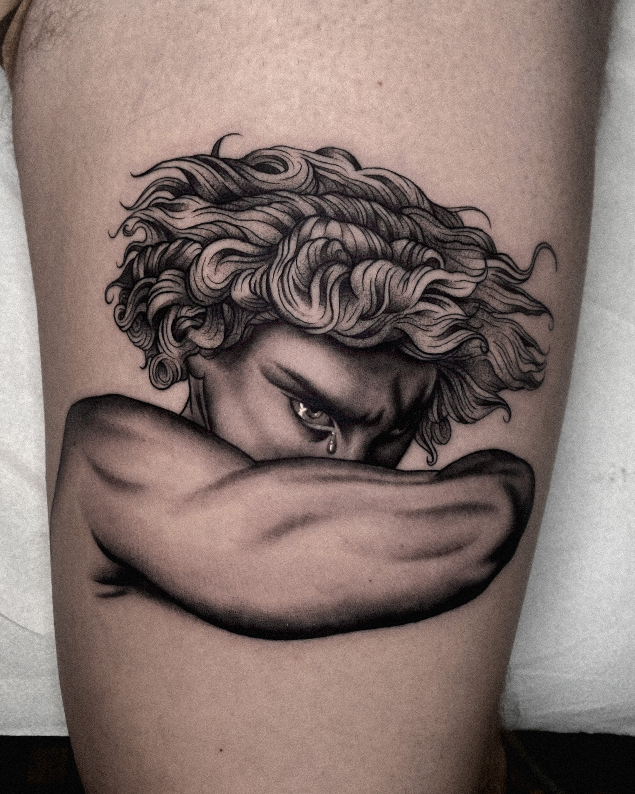Tatuaje realista de rostro en el muslo Tatuaje realista en blanco y negro de un rostro melancólico en el muslo, mostrando técnicas de sombreado y líneas finas.