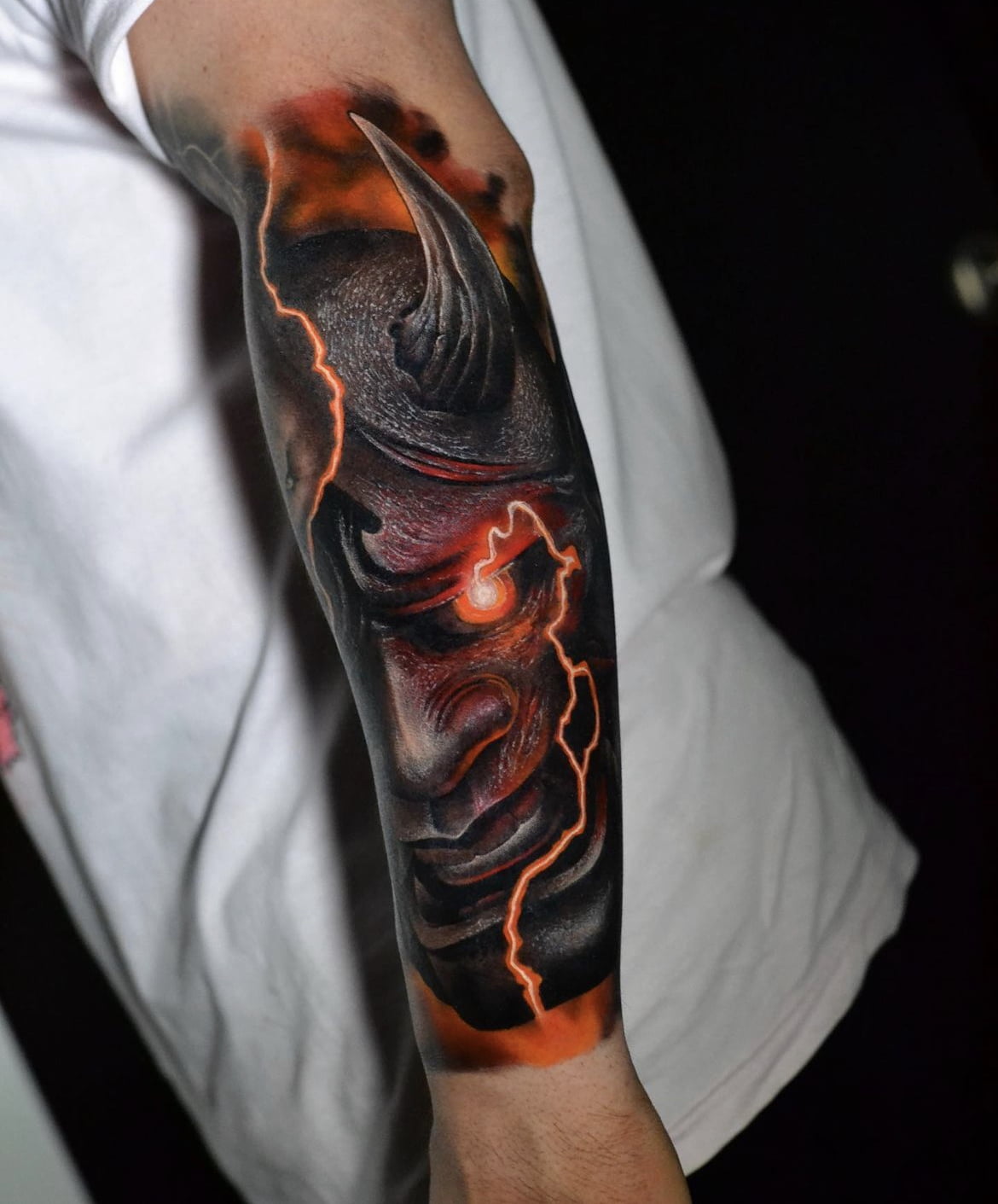 Tatuaje de máscara Oni con estilo realista en brazo, usando tonos oscuros y efectos de luz, transmitiendo una sensación de poder y misterio.