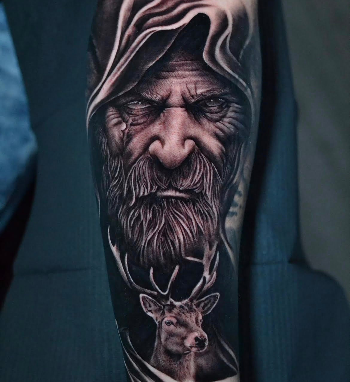 Tatuaje realista de un anciano con capucha y un ciervo en estilo black and grey en el brazo, transmitiendo sabiduría y misterio.