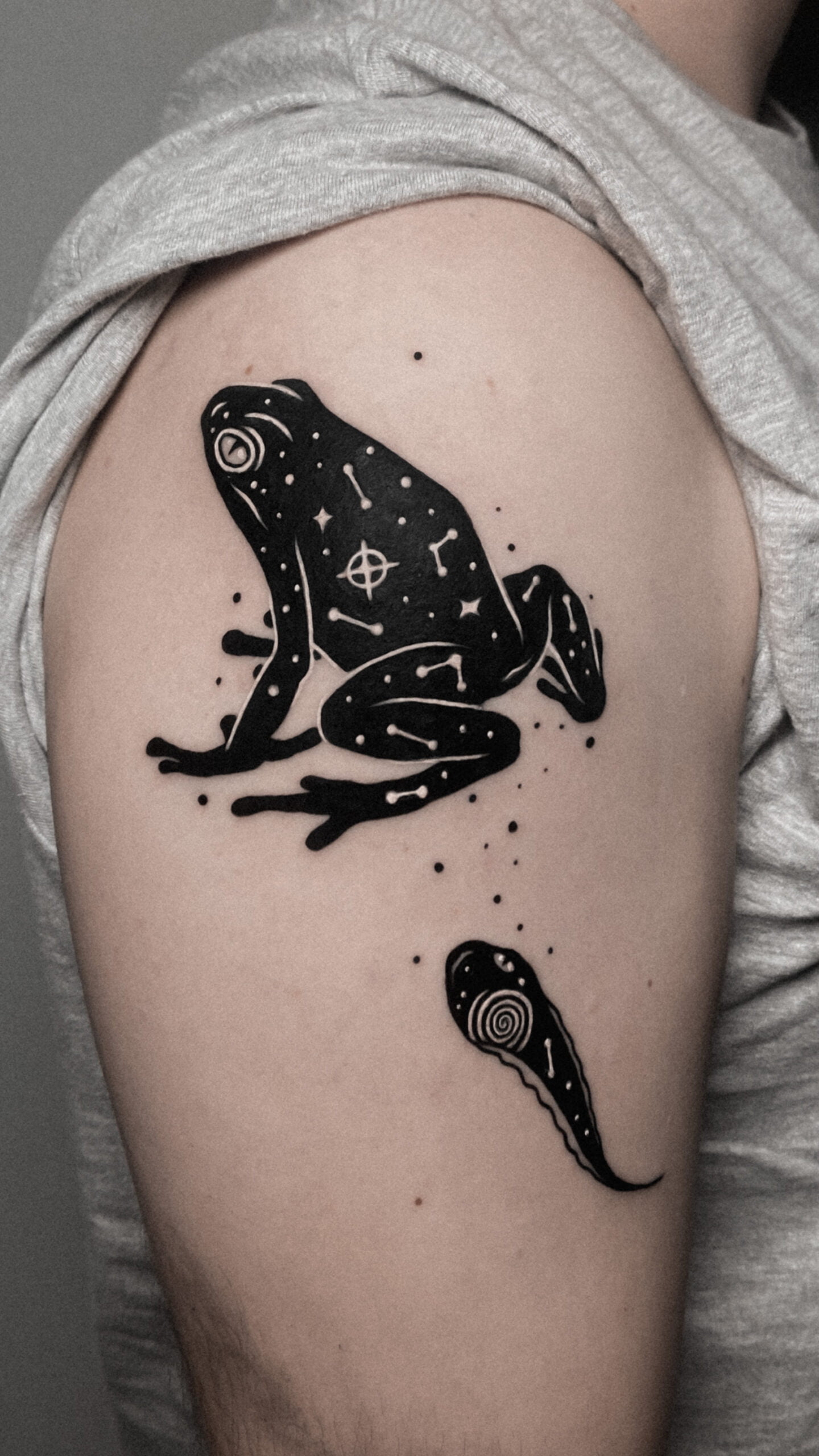 Tatuaje de rana y renacuajo en estilo blackwork en el brazo, con detalles cósmicos, transmitiendo misterio.
