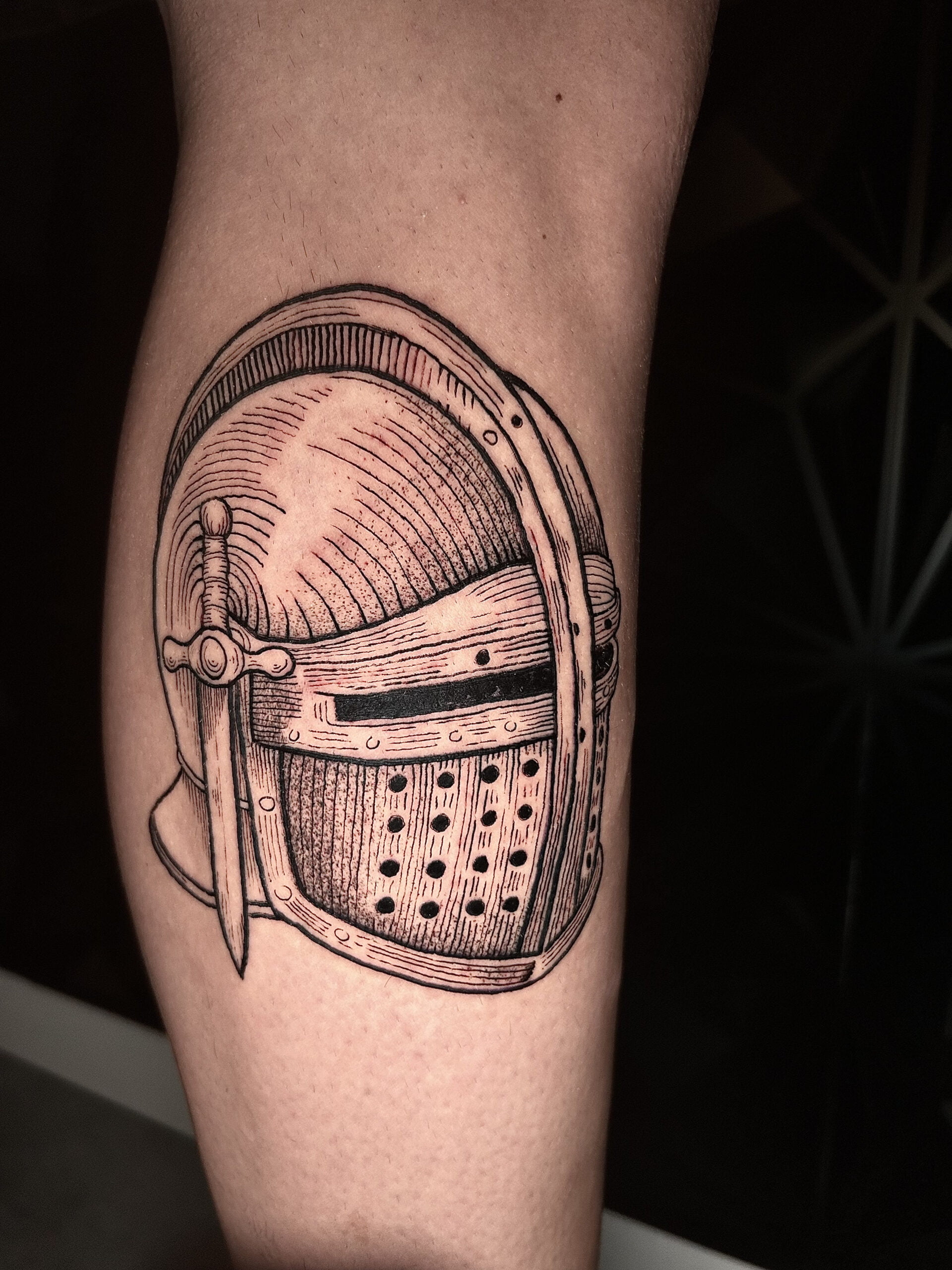 Tatuaje de casco medieval en estilo blackwork en la pierna, con líneas finas y sombreado detallado.