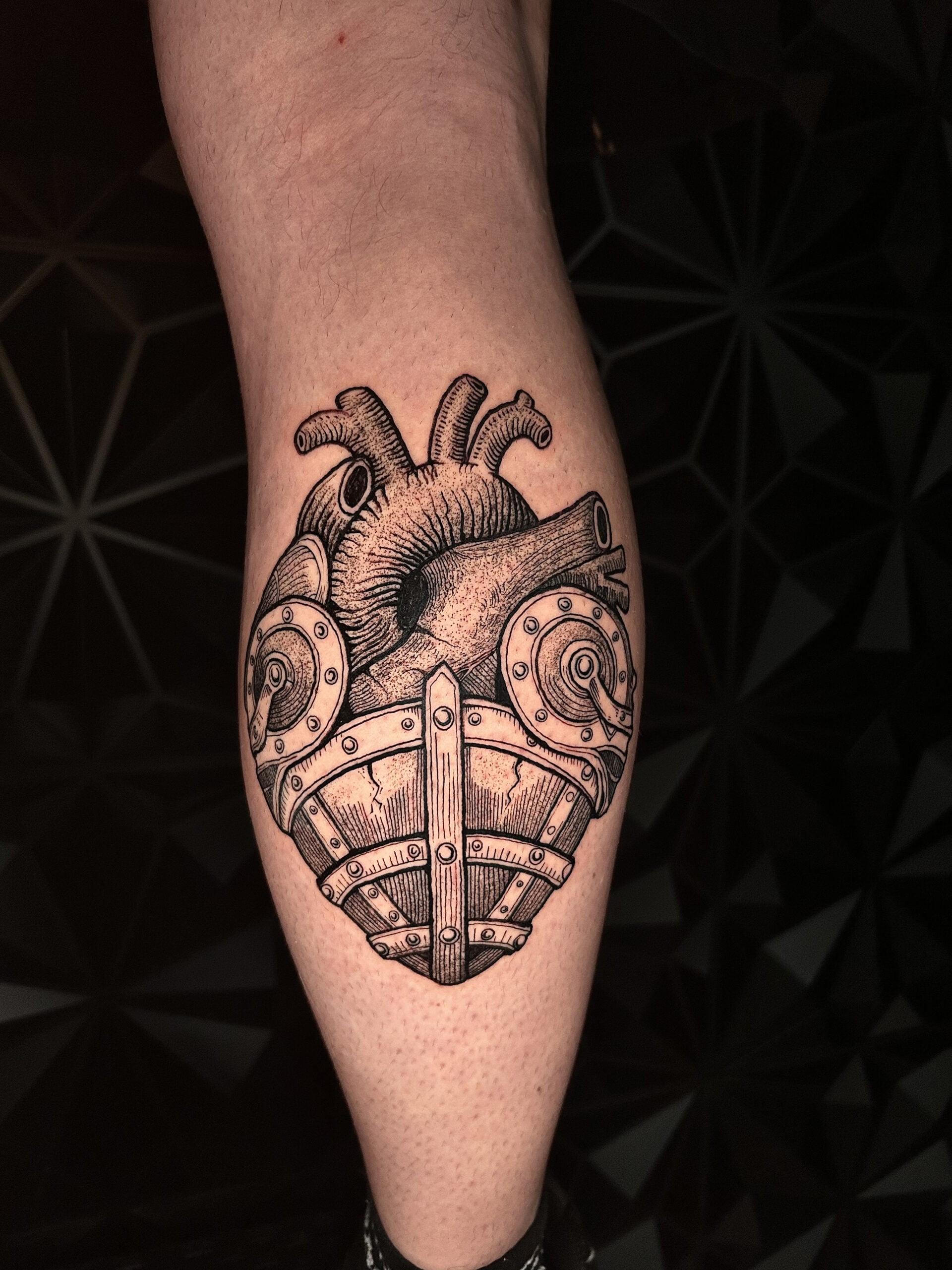 Tatuaje de un corazón anatómico protegido por una armadura en estilo blackwork en la pierna, transmitiendo fortaleza.
