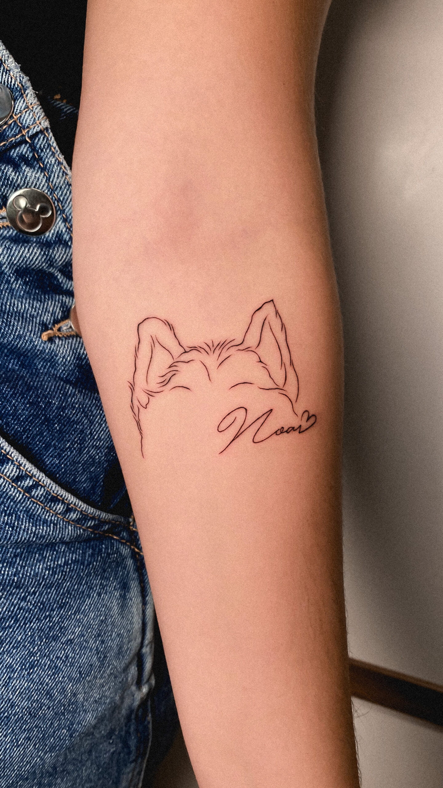 Tatuaje fineline de orejas de perro en el brazo con firma elegante y corazón.