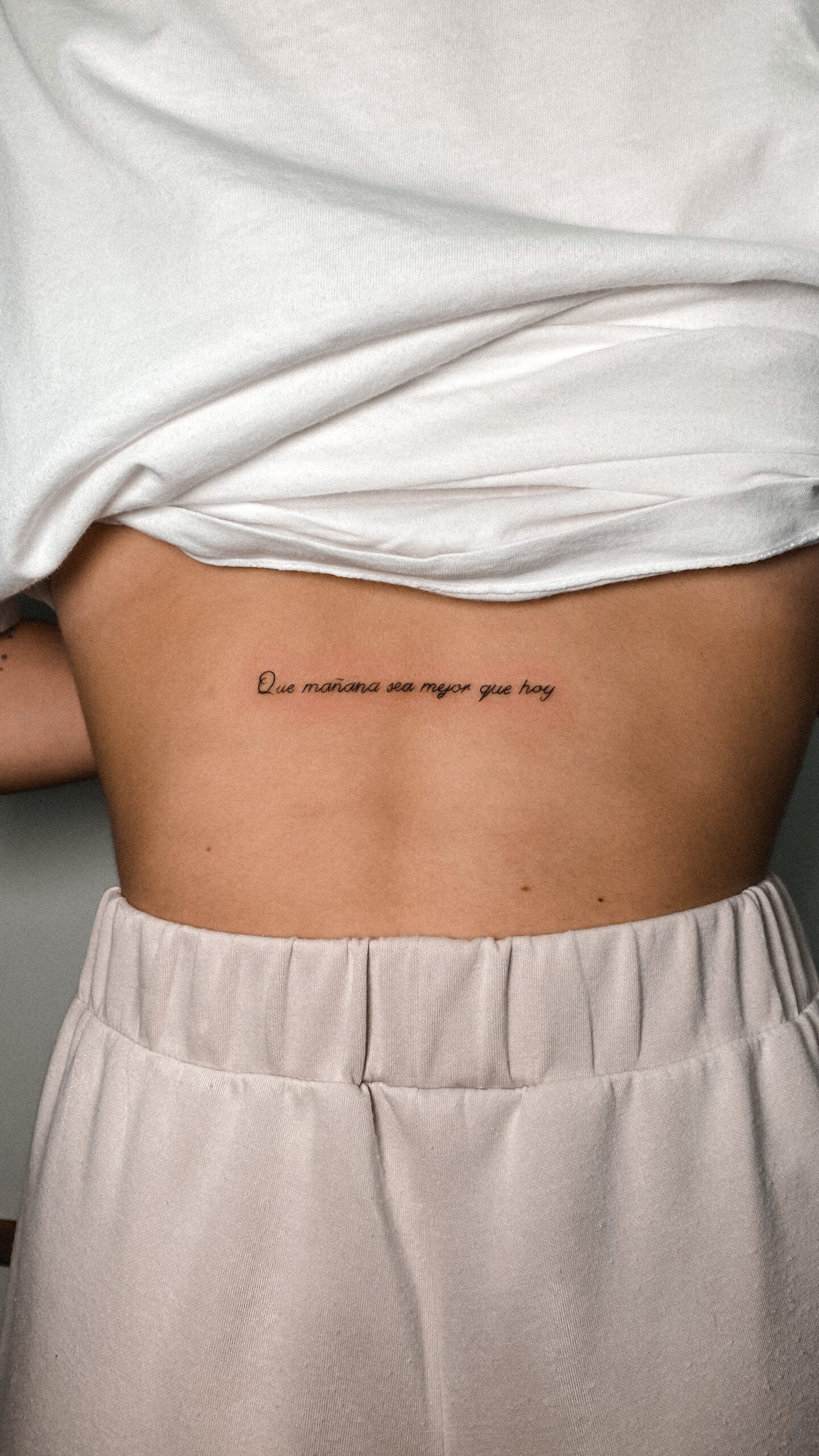 Frase motivacional en estilo fineline en la espalda baja.