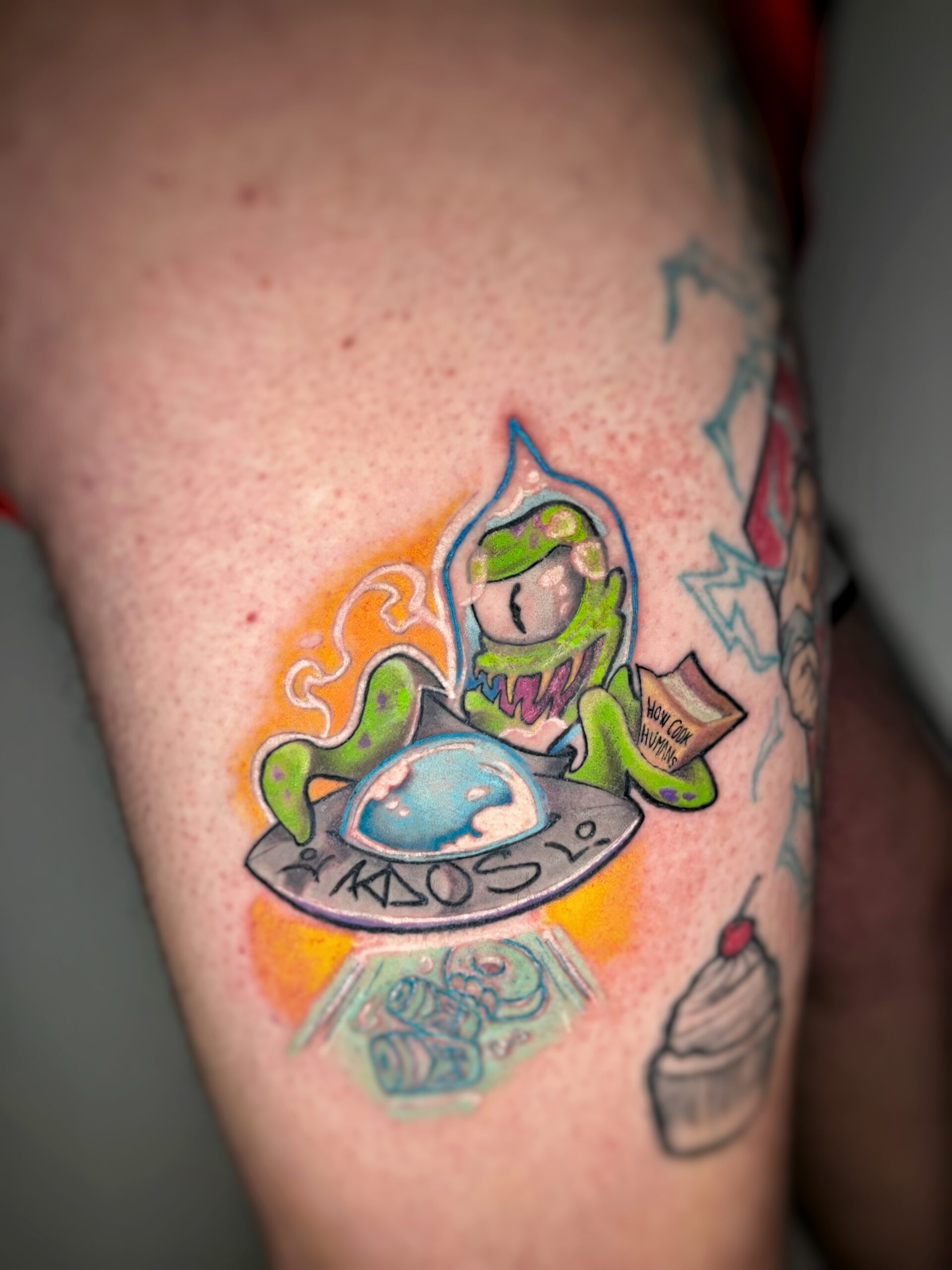 Tatuaje de un extraterrestre colorido en estilo neotradicional en el muslo, mostrando un platillo volante y un mensaje humorístico.
