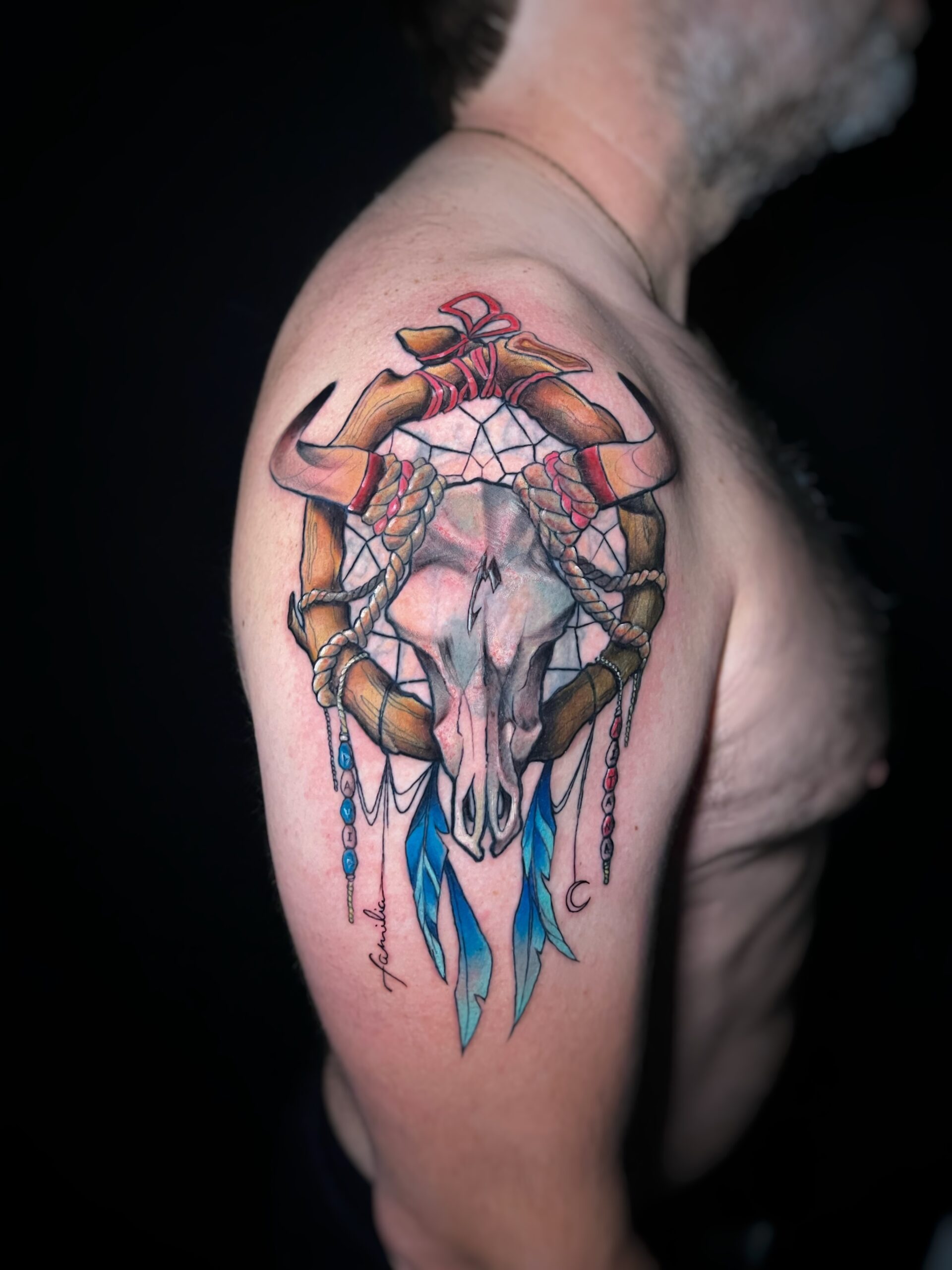 Tatuaje de cráneo de toro con atrapasueños en estilo neotradicional en el hombro, con detalles en azul y rojo.